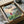 May include: Framed Ponyo movie poster print in wooden frame, packed in cardboard shipping box with protective tissue paper. Artwork shows young girl in red dress waving at seaside village with boats in harbor, rendered in Studio Ghibli's distinctive animated style with soft pastels and watercolor-like textures. Japanese text visible at top of vintage-style poster design.
