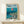 May include: Framed Studio Ghibli Ponyo poster displayed on light wooden shelf against white wall. Artwork shows red-haired Ponyo character floating in ocean waves above coastal Japanese village with traditional buildings and boats. Vibrant blues, greens, and coral colors in anime illustration style. Natural wood frame, small wrapped gift box with twine sits in front. Clean, minimalist home decor setting.