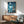 May include: Framed Studio Ghibli-style poster showing young girl with reddish hair in pink dress standing on cliff overlooking swirling ocean waves under moonlit sky with Japanese text, displayed above wooden credenza with ceramic lamp in modern living room with mint green armchair and natural lighting casting shadows on white walls.