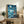 May include: Framed Studio Ghibli movie poster featuring young girl with short brown hair in red dress standing on cliff overlooking swirling blue ocean under crescent moon and starry night sky. Japanese text at top reads "Ponyo on the Cliff by the Sea." Poster displayed on rustic wooden sideboard with books, ceramic vase, and fern plant in cozy living room setting with beige armchair.