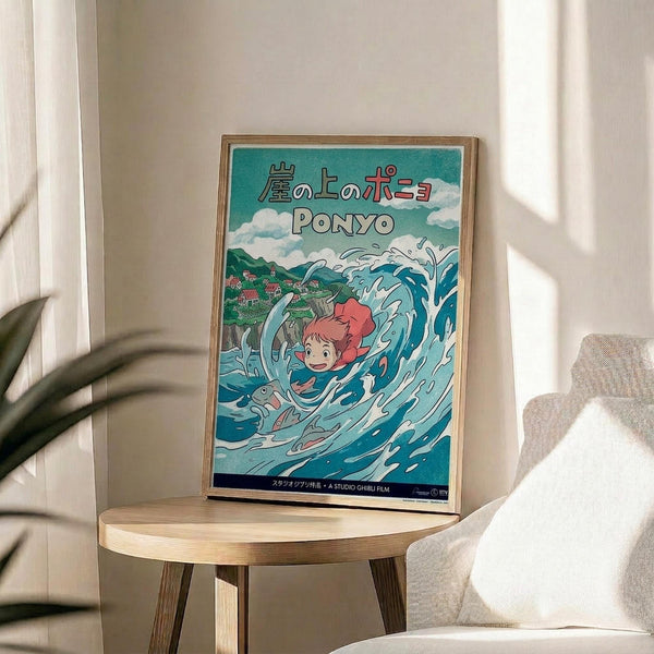 May include: A framed Studio Ghibli Ponyo movie poster displayed on a light wood round side table next to a beige upholstered chair. The colorful poster features a red-haired child character swimming in swirling blue ocean waves with fish, and a coastal village in the background. Japanese text appears at the top with "PONYO" in white letters below. Natural sunlight creates soft shadows across the neutral-toned room setting with a plant visible in the corner.