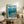 May include: Framed Studio Ghibli Ponyo poster displayed on rustic wooden sideboard in cozy living room setting. Artwork shows red-haired Ponyo character floating in turquoise ocean waters above coastal village scene. Natural wood frame, approximately 16x20 inches, positioned alongside potted fern, ceramic vases, and stacked books. Cream armchair with brown throw pillow visible in foreground against neutral wall backdrop.
