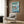 May include: Framed Ponyo poster displayed on wood-paneled wall above beige upholstered chair with geometric pillow. Poster shows red-haired character riding ocean wave in anime art style with Japanese text and turquoise background. Modern room setting with brass floor lamp and round side table.