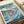 May include: Framed Studio Ghibli Ponyo poster print displayed on white bedding next to a cream pillow. The artwork shows red-haired Ponyo character surrounded by swirling blue ocean waves with a coastal village in the background. Japanese text appears at the top, with "Studio Ghibli Film" credited at bottom. Print has a light wooden frame and vintage-style illustration in teal, coral, and white tones.