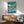 May include: A framed Studio Ghibli Ponyo poster displayed on a white wall above a mid-century modern wooden credenza. The poster features the red-haired character Ponyo swimming in blue ocean waves with a fish, set against a turquoise background with Japanese text. A teal upholstered armchair with wooden frame sits nearby alongside a small table lamp and round coffee table.