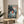 May include: Framed Studio Ghibli Ponyo poster displayed on light wood round side table next to beige upholstered chair. Artwork shows orange-haired child character splashing in ocean waves between rocky coastal cliffs, with Japanese and English text at top. Natural sunlight streams across white walls, with green plant leaves visible in corner.