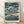 May include: Framed poster print in light wood frame leaning against white textured wall on wooden floor. Artwork shows animated scene with two children floating in stormy ocean waves, submarine visible, dramatic cloudy sky above coastal houses and boats. Japanese text and English subtitle at bottom. Poster appears medium-sized with white matting around colorful illustration in Studio Ghibli anime style.