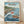 May include: Framed poster displaying Studio Ghibli-style artwork of Ponyo on the Cliff by the Sea. Shows red-haired girl character surfing on a large blue wave next to a wooden fishing boat with a boy aboard. Background features coastal Japanese village with lighthouse, rocky cliffs, and green hills. Japanese and English text overlay. Light wood frame against white wall on wooden floor with soft natural lighting.