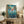 May include: Framed Studio Ghibli Ponyo movie poster displayed on rustic wooden sideboard next to cream armchair, fern plant, and ceramic vase. Colorful anime artwork shows red-haired character emerging from swirling ocean waters with coastal village and lighthouse in background. Japanese text visible with English subtitle. Warm, cozy living room setting with natural lighting and earthy tones throughout the staged scene.