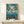 May include: Framed Studio Ghibli Ponyo movie poster displayed on light wood console table with small wrapped gift box. Poster shows red-haired Ponyo character emerging from swirling ocean waves with coastal town, lighthouse, and underwater sea life including fish and jellyfish. Artwork features vibrant blues, greens, and coral tones in classic anime illustration style with Japanese text and thin wooden frame.