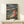May include: Framed Studio Ghibli Ponyo movie poster displayed on light wood shelf against cream wall. Poster shows red-haired child in red dress sitting on rocky coastline holding jar, with turbulent ocean waves and clifftop village in background under stormy sky. Japanese and English text at top. Small white wrapped gift box sits in front of frame on shelf.