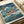 May include: Studio Ghibli Ponyo poster print displayed on white paper with cream ribbon nearby. The colorful anime artwork shows characters in turbulent ocean waves with boats and houses in background. Japanese and English text at bottom reads "Ponyo on the Cliff by the Sea." Vintage-style illustration features blue-green stormy seas, red-haired girl character, and boy in yellow jacket amid dramatic white-capped waves.