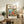 May include: Framed Studio Ghibli poster featuring Ponyo character in red dress cooking in cozy kitchen overlooking ocean cliffs, displayed on rustic wooden sideboard with potted fern, ceramic vase, and stack of books in warm, sunlit living room with cream upholstered furniture and neutral walls.