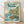 May include: Framed Studio Ghibli Ponyo poster in light wood frame, leaning against white wall on wooden floor. Artwork shows young girl with reddish hair in orange dress standing on rocky outcrop surrounded by ocean waves, with cliffside village houses visible in background. Japanese text at top, English subtitle reads "A magical journey begins where the sea meets the shore." Soft natural lighting creates gentle shadows on white textured wall.
