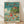 May include: Framed poster print in light wood frame leaning against white wall on wooden floor. Features Studio Ghibli anime-style artwork showing children swimming and playing in blue-green ocean water with fish, while a house sits on clifftop surrounded by lush green trees under cloudy sky. Japanese text at top with "Ponyo" subtitle. Vibrant illustration in classic animation poster style with turquoise, green, and warm earth tones.