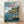 May include: Framed poster print leaning against white wall on light wooden floor. The artwork shows Studio Ghibli's Ponyo anime scene with green-haired boy sitting on wooden dock, red-haired girl swimming in blue water below. Japanese coastal village with traditional houses built into rocky cliffs visible in background under cloudy sky. Japanese text at top in beige and red lettering. Natural wood frame surrounds the colorful illustration in signature Miyazaki animation style.