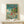 May include: Framed Studio Ghibli Ponyo poster displayed on light wood shelf against white wall. Artwork shows children swimming in turquoise ocean water with fish, featuring red-haired Ponyo character prominently. House on cliff visible in background with blue sky and white clouds. Poster has thin natural wood frame. Small white wrapped gift box sits on shelf in front of poster. Clean, minimalist room setting with natural lighting casting soft shadows.