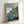 May include: Framed Studio Ghibli Ponyo movie poster print leaning against white wall on light surface. Colorful anime-style artwork shows seaside cliff village with traditional Japanese houses, blue ocean with swimming red-haired girl character, and green-haired child on wooden dock. Japanese text and "PONYO" title at top against cloudy sky background. Natural wood frame surrounds the vibrant illustration.