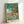 May include: Framed poster print featuring animated children playing in ocean waves with fish swimming nearby, house on cliff in background under blue sky with white clouds. Japanese text at top reads "Ponyo" in golden letters. Artwork displays Studio Ghibli animation style with vibrant blues, greens, and warm earth tones. Light wood frame leans against white wall in bright room setting.