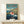 May include: Framed Studio Ghibli poster showing Ponyo, a red-haired fish girl character, swimming in dark blue ocean water near a rocky coastal village. The artwork features Japanese text at top with English subtitle "Ponyo on the Cliff by the Sea." Natural wood frame sits on light wooden shelf against white wall, with small wrapped gift box below.