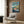 May include: Framed Ponyo on the Cliff by the Sea movie poster displayed on wood-paneled wall above beige armchair with geometric pillow. Poster shows red-haired child character swimming in blue ocean water near coastal houses on rocky cliffs. Modern room setting with floor lamp and side table.