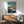 May include: Framed Ponyo on the Cliff by the Sea movie poster displayed above a mid-century modern wooden credenza with table lamp. The poster features the red-haired character Ponyo splashing in blue water with a coastal village backdrop. Room has white walls, natural lighting, and a teal upholstered chair with wooden arms visible in corner.
