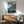 May include: Framed Ponyo poster on white wall above wooden credenza with lamp and mid-century modern teal armchair. Poster shows animated scene with boy on wooden dock, red-haired girl swimming in blue water, and coastal village with traditional houses on clifftop. Japanese text and "PONYO" title visible. Warm natural lighting creates geometric shadows across minimalist room setting.