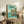 May include: Framed Studio Ghibli Ponyo poster displayed on rustic wooden credenza in living room setting. Poster features young girl in red dress standing on rocky outcrop surrounded by ocean waves, with Japanese text and coastal cliff scenery in soft blues and greens. Room includes fern plant, ceramic vase, books, and beige upholstered furniture with brown throw pillow.