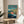 May include: Framed poster of Studio Ghibli's Ponyo displayed on a round wooden side table next to a beige upholstered chair. The colorful anime-style artwork shows a red-haired girl character swimming in blue ocean water near a coastal cliff village. Japanese and English text appears at the top against a turquoise sky with white clouds. Natural lighting creates soft shadows across the minimalist room setting with white walls and a green plant visible in the corner.