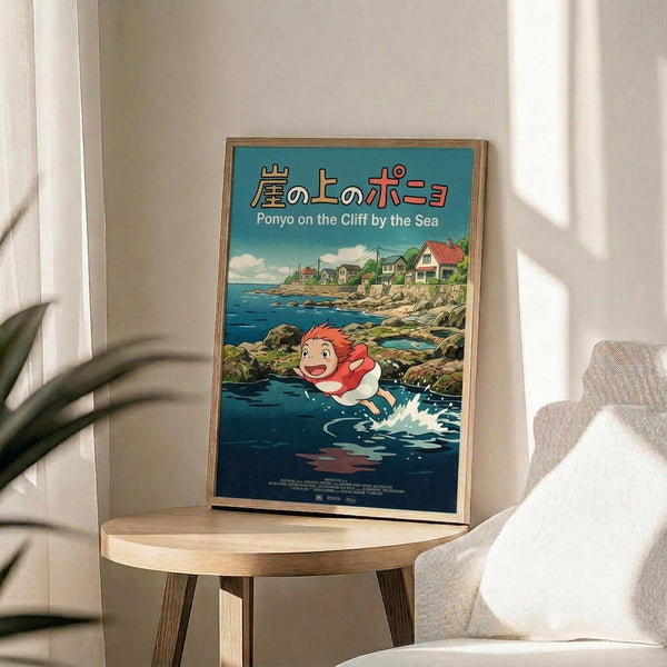 May include: Framed poster of Studio Ghibli's Ponyo displayed on a round wooden side table next to a beige upholstered chair. The colorful anime-style artwork shows a red-haired girl character swimming in blue ocean water near a coastal cliff village. Japanese and English text appears at the top against a turquoise sky with white clouds. Natural lighting creates soft shadows across the minimalist room setting with white walls and a green plant visible in the corner.