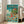 May include: A framed poster featuring Studio Ghibli's Ponyo in vibrant anime style, showing children swimming in turquoise ocean water with colorful fish, a clifftop house with red roof surrounded by green trees against blue sky with white clouds, Japanese text at top, displayed in bright living room setting with beige sofa and large windows.