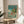 May include: Framed Studio Ghibli Ponyo movie poster displayed on wooden side table next to beige upholstered chair. Poster shows colorful anime illustration of children swimming in ocean waves with fish, house on cliff above, Japanese text at top. Natural wood frame, bright turquoise and coral colors, sunlight streaming across white walls and plant leaves visible in corner.