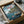 May include: Framed Ponyo poster print partially unwrapped in cardboard shipping box with white tissue paper. Artwork shows anime characters by waterfront village - boy on wooden dock, small red-haired character in blue water. Coastal scene with traditional houses on cliffs, green trees, cloudy sky. Japanese text at top. Protective brown cardboard corners visible on black frame.
