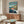May include: Studio Ghibli-style poster mounted on beige wall above wooden credenza in modern living room. Poster shows red-haired boy swimming in blue ocean water near coastal cliffs with houses, featuring Japanese and English text. Room has white curved chair, glass coffee table, floor lamp, and minimalist mid-century decor with warm wood tones and neutral colors.