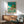 May include: Framed Ponyo movie poster displayed above a wooden sideboard in a modern living room. The poster shows colorful anime-style illustration with children swimming in turquoise water near a clifftop house. Features teal background, Japanese text, and Studio Ghibli artwork style. Room has mid-century furniture, gray upholstered chair, white table lamp, and natural lighting casting shadows on white walls.