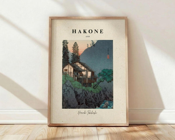 May include: Framed vintage Japanese woodblock print showing traditional houses nestled among green foliage and rocky terrain, with mountains in background and peachy sunset sky. Light wood frame with cream matting, leaning against white wall on natural wood floor with soft window light creating diagonal shadows.