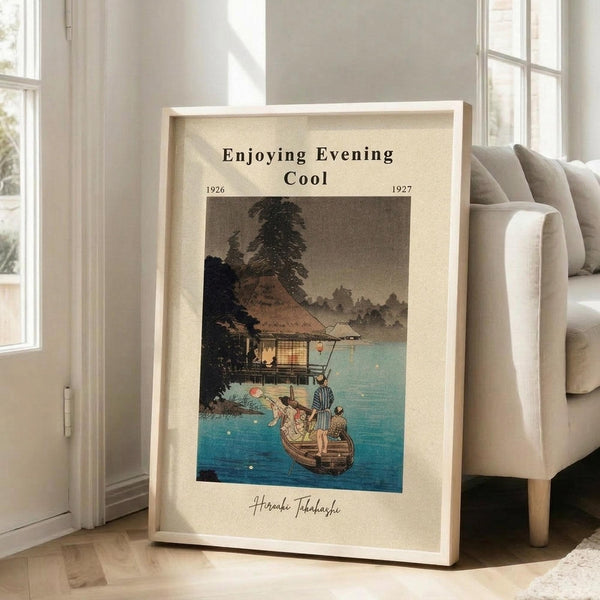 May include: Framed vintage Japanese woodblock print showing people in a boat near a traditional thatched-roof house by water at dusk. The artwork features muted blues, browns, and greens with "Enjoying Evening Cool" text and dates 1926-1927. The beige-matted print in light wood frame leans against white wall beside cream upholstered chair in bright, minimalist living room with large windows.