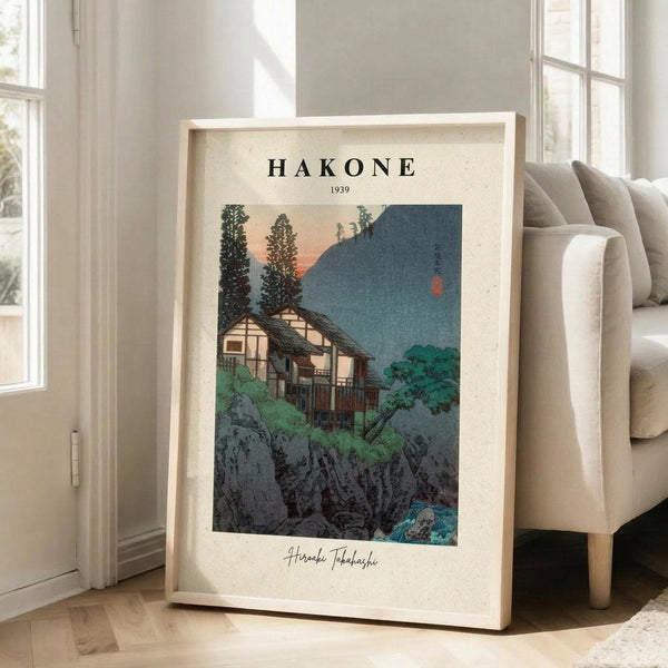 May include: Framed vintage-style Japanese woodblock print poster showing traditional houses nestled among rocks and trees with mountains in background, displaying muted greens, blues, and warm sunset tones. Light wood frame leans against white wall beside beige upholstered chair in bright, minimalist living room with large windows and hardwood floors.