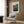 May include: Vintage Japanese travel poster featuring traditional buildings nestled among mountains and trees in muted blue, green, and coral tones, displayed in cream matting and light wood frame on wood-paneled wall above beige upholstered armchair with geometric patterned throw pillow, alongside brass floor lamp and dark round side table with open book.