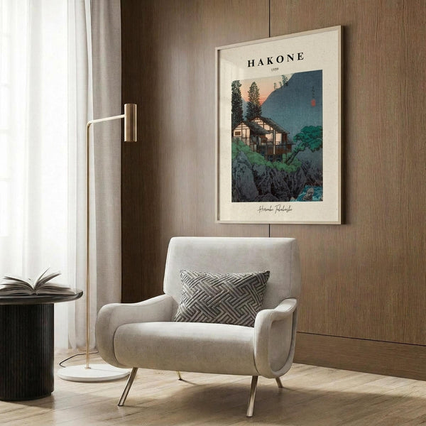 May include: Vintage Japanese travel poster featuring traditional buildings nestled among mountains and trees in muted blue, green, and coral tones, displayed in cream matting and light wood frame on wood-paneled wall above beige upholstered armchair with geometric patterned throw pillow, alongside brass floor lamp and dark round side table with open book.