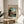 May include: Framed vintage-style Japanese woodblock print showing Mount Fuji in distance with autumn foliage, rocky stream, and traditional landscape elements in muted blues, golds, and greens. Print sits on round wooden side table next to beige upholstered chair with plant leaves visible, soft natural lighting creating shadows on white wall.