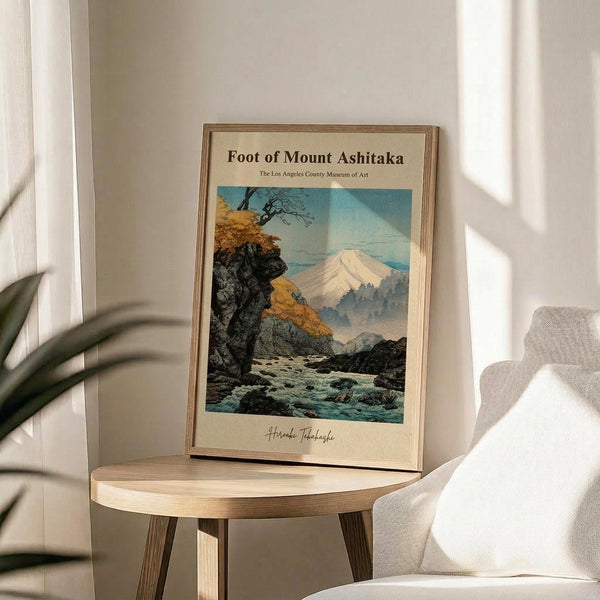 May include: Framed vintage-style Japanese woodblock print showing Mount Fuji in distance with autumn foliage, rocky stream, and traditional landscape elements in muted blues, golds, and greens. Print sits on round wooden side table next to beige upholstered chair with plant leaves visible, soft natural lighting creating shadows on white wall.