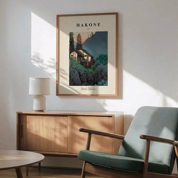 May include: Framed vintage Japanese woodblock print showing traditional buildings nestled among trees and rocky terrain with mountains in background, displayed above wooden sideboard with lamp in modern living room. Mid-century green armchair with wooden arms sits nearby. Warm natural lighting creates diagonal shadows across white wall. Print features muted greens, blues, and earth tones in classic ukiyo-e style.