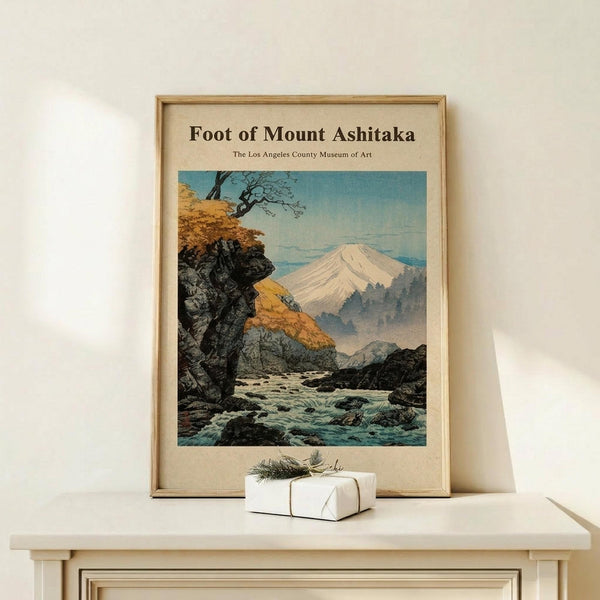 May include: Framed vintage-style Japanese woodblock print showing Mount Fuji in background with snow-capped peak, rocky stream with flowing water in foreground, autumn trees with golden foliage on left side. Natural wood frame on light wooden console table against cream wall, small white wrapped gift with twine and pine sprig positioned in front.