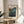 May include: Framed vintage Japanese woodblock print poster showing traditional houses nestled among evergreen trees and mountains at dusk, with blue peaks and orange sky. The cream-colored print displays "HAKONE 1939" text and artist signature. Displayed on round wooden side table next to beige upholstered chair, with plant leaves visible in corner and natural sunlight casting shadows on white wall.