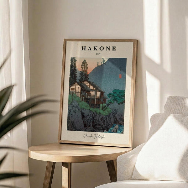 May include: Framed vintage Japanese woodblock print poster showing traditional houses nestled among evergreen trees and mountains at dusk, with blue peaks and orange sky. The cream-colored print displays "HAKONE 1939" text and artist signature. Displayed on round wooden side table next to beige upholstered chair, with plant leaves visible in corner and natural sunlight casting shadows on white wall.