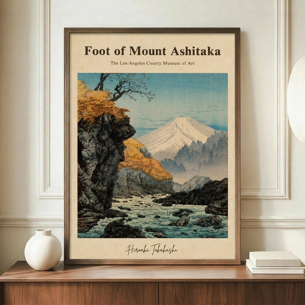May include: Framed vintage Japanese woodblock print showing Mount Fuji with snow-capped peak in background, rocky stream with flowing water in foreground, autumn foliage on cliffs, displayed on wooden credenza next to white ceramic vase and stacked books against white paneled wall