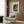 May include: Framed vintage Japanese woodblock print poster showing evening lakeside scene with traditional house, boat, and figures in blue and earth tones, displayed on wood paneled wall above beige upholstered armchair with geometric patterned pillow, alongside brass floor lamp and dark side table with open book