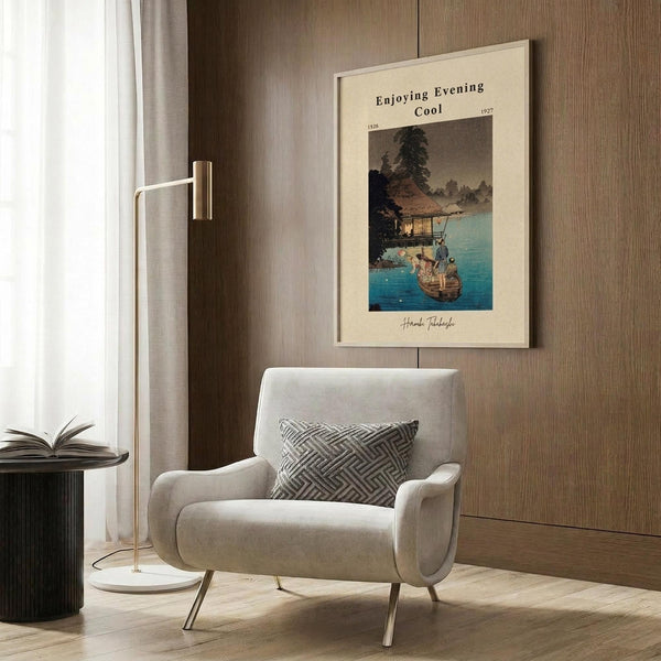 May include: Framed vintage Japanese woodblock print poster showing evening lakeside scene with traditional house, boat, and figures in blue and earth tones, displayed on wood paneled wall above beige upholstered armchair with geometric patterned pillow, alongside brass floor lamp and dark side table with open book