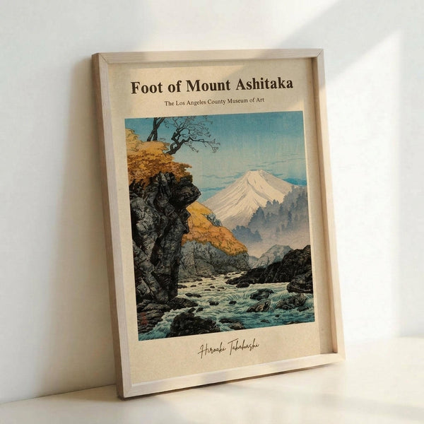 May include: Framed vintage Japanese woodblock print showing Mount Fuji in background with autumn foliage, rocky cliffs, and flowing stream in foreground. Print features warm oranges, blues, and earth tones in traditional ukiyo-e style. Light wood frame sits against white wall, displaying museum poster with artist signature at bottom.